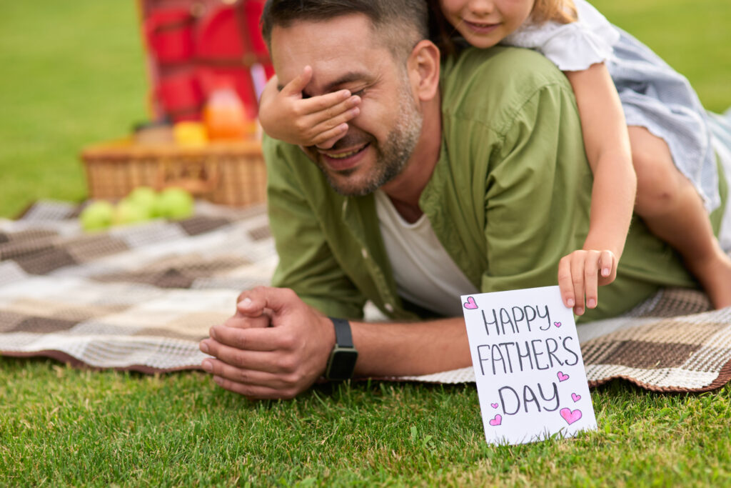 Cute little girl surprising her father, greeting and giving him handmade postcard, daddy and daughter celebrating Father's Day in the park